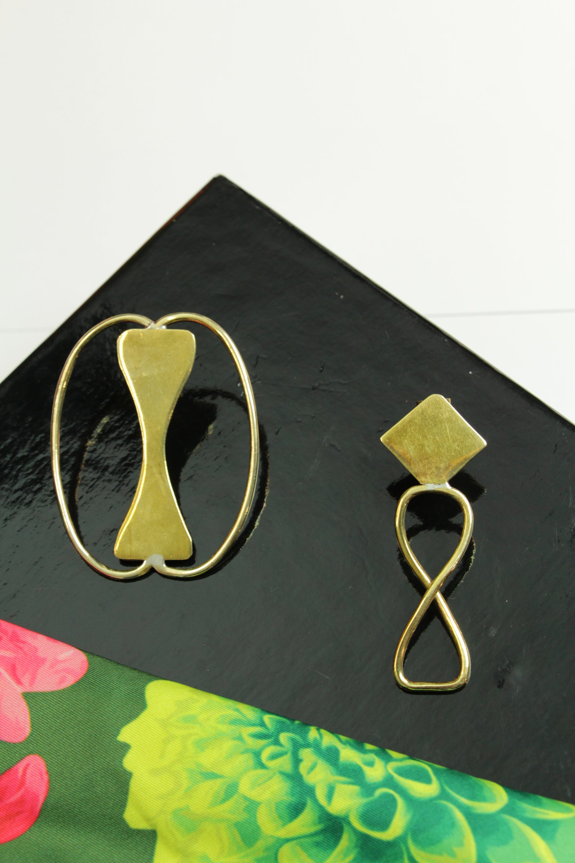 Boucles d'oreilles Adna posées sur un socle noir près d'un foulard vert imprimé feuilles et fleurs